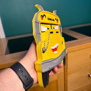 K&ouml;nnte beinhalten: Ein gelbes Schulbus-Spielzeug mit einem l&auml;chelnden Gesicht, schwarzen Reifen und silbernen Akzenten. Der Bus tr&auml;gt die Aufschrift "SCHOOL BUS" oben. Das Spielzeug wird in der Hand einer Person gehalten.