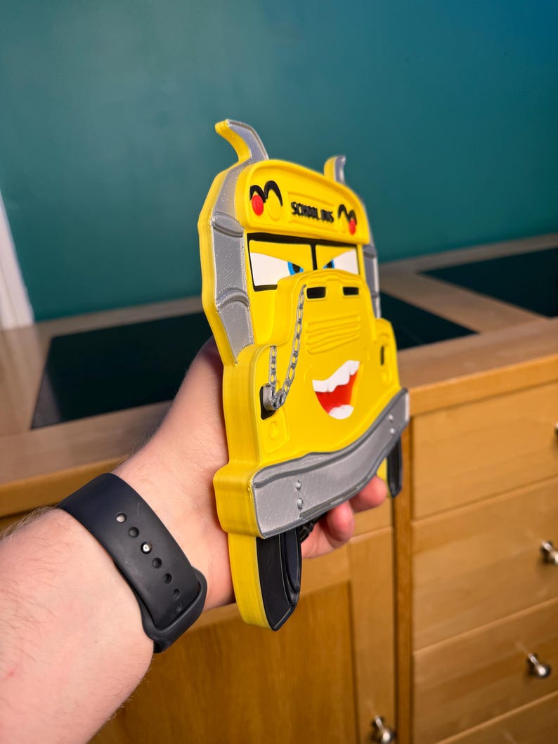 K&ouml;nnte beinhalten: Ein gelbes Schulbus-Spielzeug mit einem l&auml;chelnden Gesicht, schwarzen Reifen und silbernen Akzenten. Der Bus tr&auml;gt die Aufschrift "SCHOOL BUS" oben. Das Spielzeug wird in der Hand einer Person gehalten.