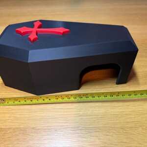 A black 3D-printed gothic reptile hide with a red cross on top, placed on a wooden surface. A measuring tape is extended alongside, showing its approximate length of 9 inches. Ideal for snakes, geckos, and other small reptiles.