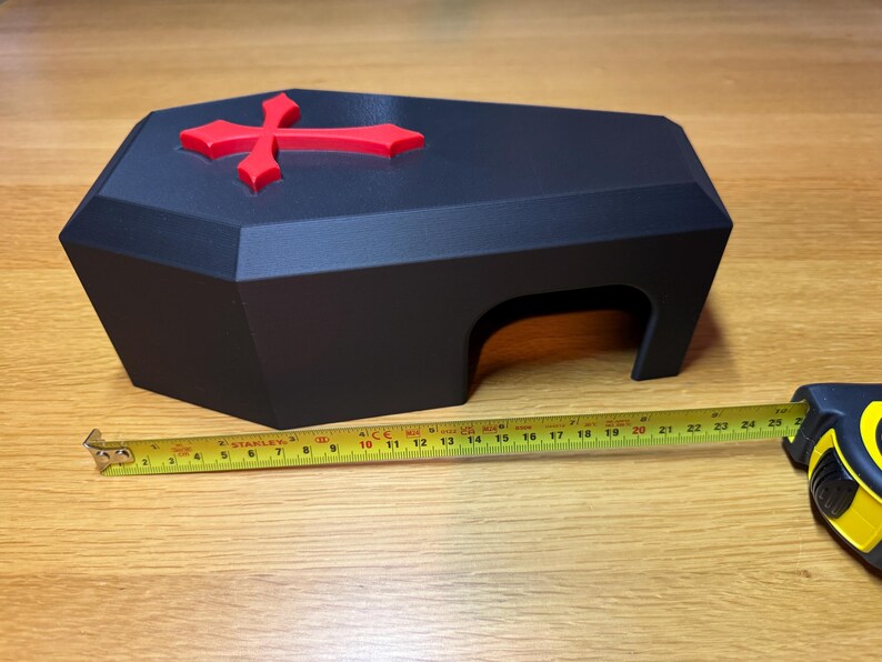 A black 3D-printed gothic reptile hide with a red cross on top, placed on a wooden surface. A measuring tape is extended alongside, showing its approximate length of 9 inches. Ideal for snakes, geckos, and other small reptiles.