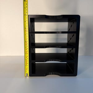 May include: A black, four-tiered shelf unit with curved edges. The unit is approximately 13 inches tall, as measured by a yellow tape measure. The shelves are evenly spaced and appear to be made of a solid material.