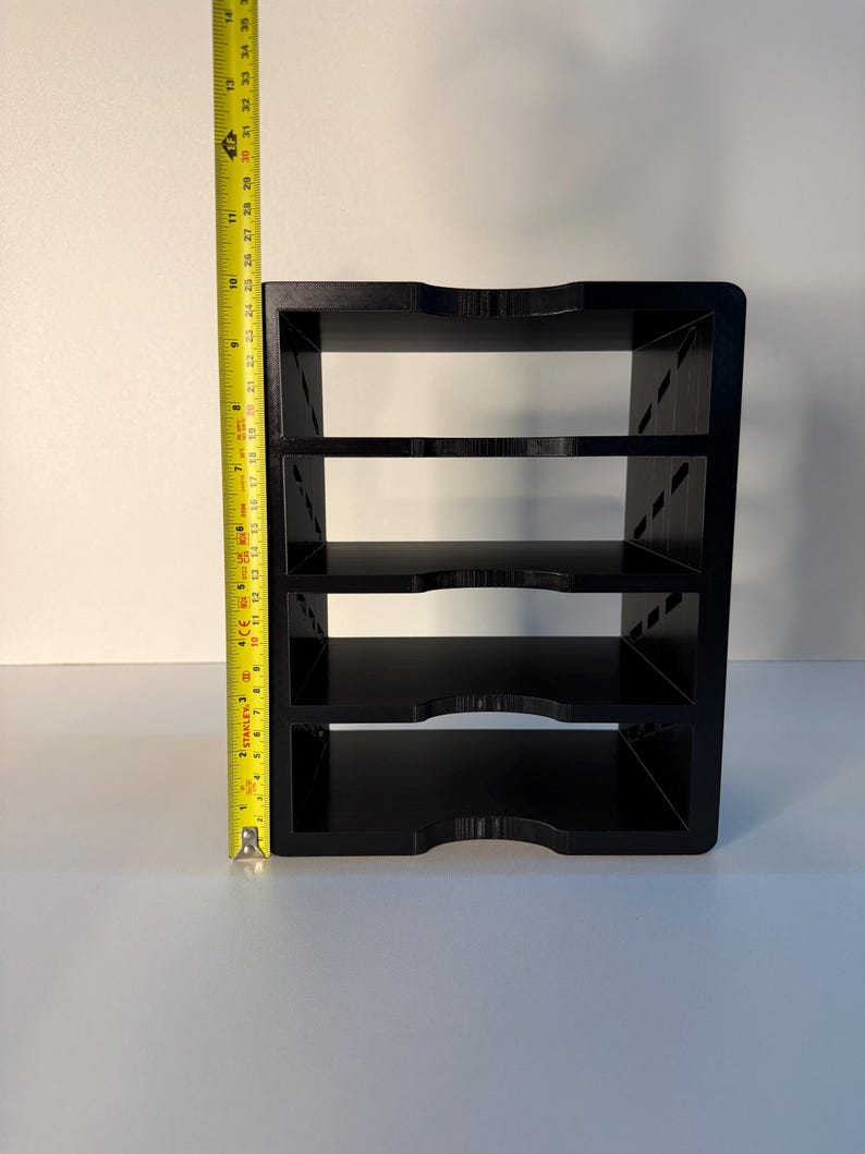 May include: A black, four-tiered shelf unit with curved edges. The unit is approximately 13 inches tall, as measured by a yellow tape measure. The shelves are evenly spaced and appear to be made of a solid material.