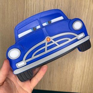 Pode incluir: Um brinquedo azul em forma de carro com detalhes brancos, far&oacute;is e olhos de desenho animado. O brinquedo tem uma grelha prateada e um emblema vermelho. As rodas s&atilde;o pretas. O brinquedo &eacute; segurado em uma m&atilde;o.