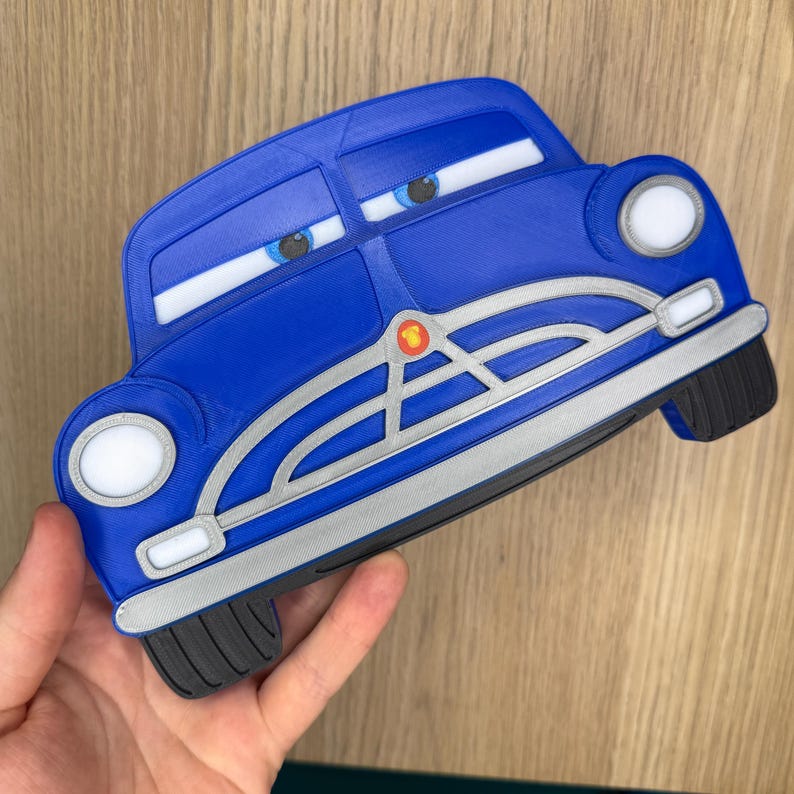 Pode incluir: Um brinquedo azul em forma de carro com detalhes brancos, far&oacute;is e olhos de desenho animado. O brinquedo tem uma grelha prateada e um emblema vermelho. As rodas s&atilde;o pretas. O brinquedo &eacute; segurado em uma m&atilde;o.