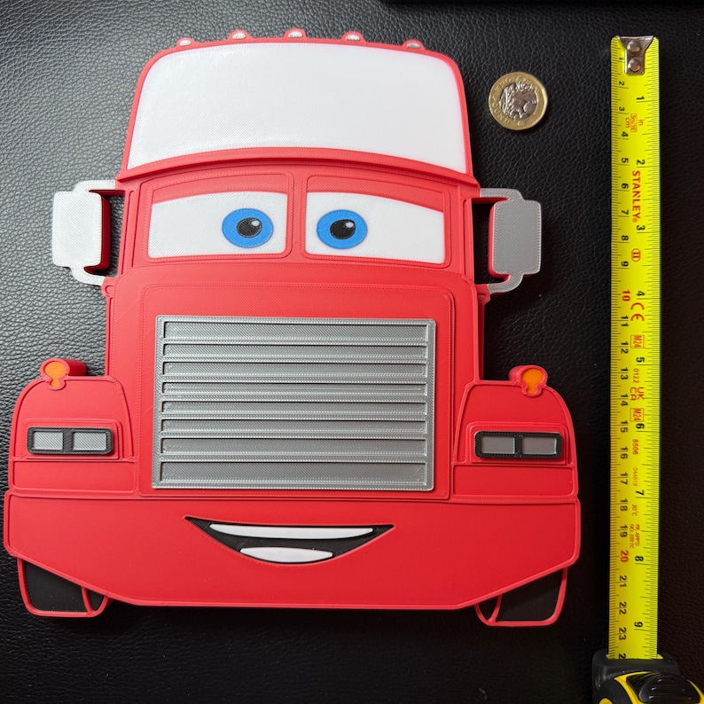 May include: A red cartoon truck-shaped object with a white top, blue eyes, and a smiling mouth. The truck has a silver grill and side mirrors. A yellow tape measure shows the truck is approximately 9 inches tall.