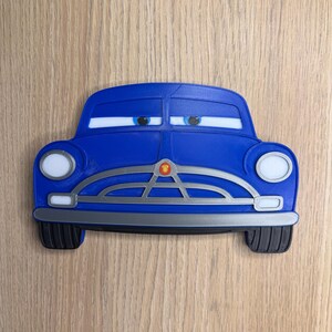 Pode incluir: Decora&ccedil;&atilde;o azul em forma de carro com um design de desenho animado. O carro tem far&oacute;is brancos, uma grelha prateada e pneus pretos. Os olhos do carro s&atilde;o azuis e tem um emblema vermelho na frente.