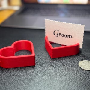 Red 3D-printed heart-shaped place card holders displayed with a “Groom” card and a coin for size comparison. Ideal for weddings, receptions, and bridal showers, these durable holders combine practicality and style.