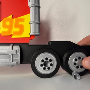 May include: A red and yellow toy truck cab with the number 95 in orange. The truck has black wheels with silver rims. The image shows a hand holding the wheels.