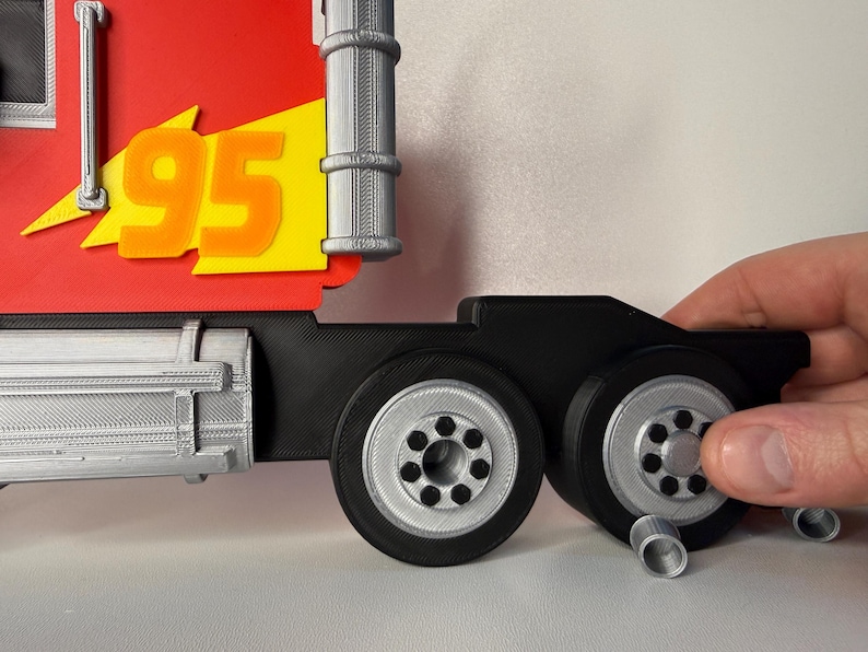 May include: A red and yellow toy truck cab with the number 95 in orange. The truck has black wheels with silver rims. The image shows a hand holding the wheels.