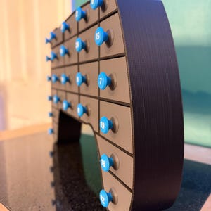 May include: A black and gray advent calendar with blue knobs. Each knob has a white number on it. The calendar has multiple drawers, each with a blue knob. The calendar is on a black surface.