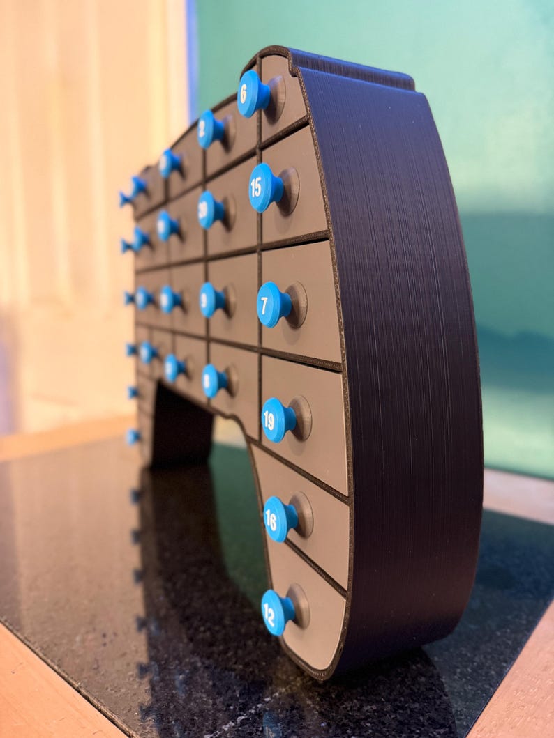 May include: A black and gray advent calendar with blue knobs. Each knob has a white number on it. The calendar has multiple drawers, each with a blue knob. The calendar is on a black surface.