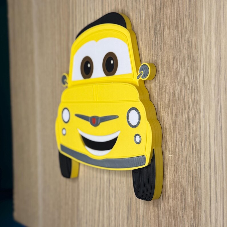 May include: A yellow cartoon car-shaped decoration with black tires and a black top. The car has a smiling face with brown eyes and a gray bumper. The decoration is mounted on a wooden surface.