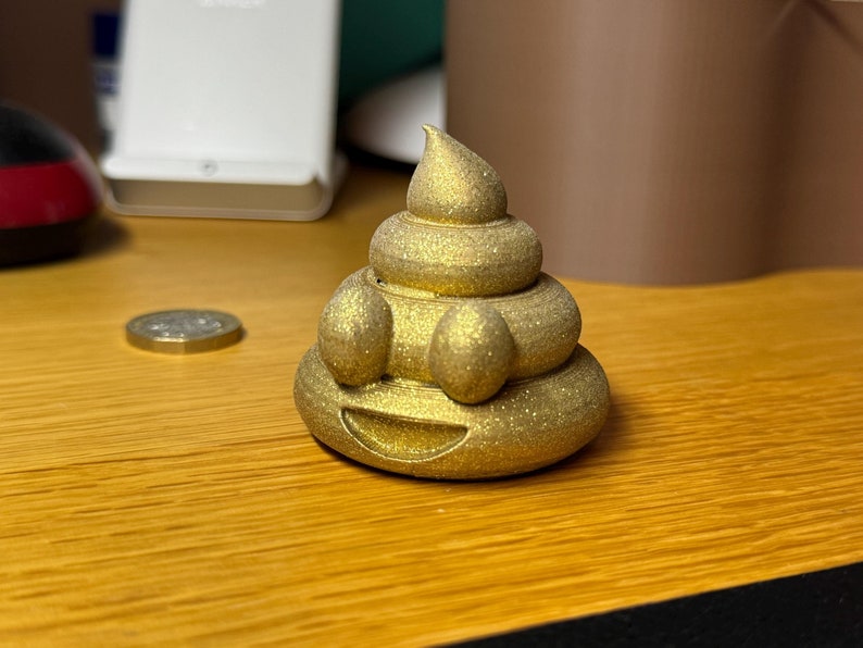 A glittery gold 3D-printed poop emoji made from recycled plastic waste, sitting on a wooden surface with a 1 pound coin in the background for size reference.