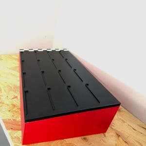 May include: A rectangular display stand with a red base and a black top. The top features several evenly spaced, vertical slots. The stand is angled, showcasing its design and construction.