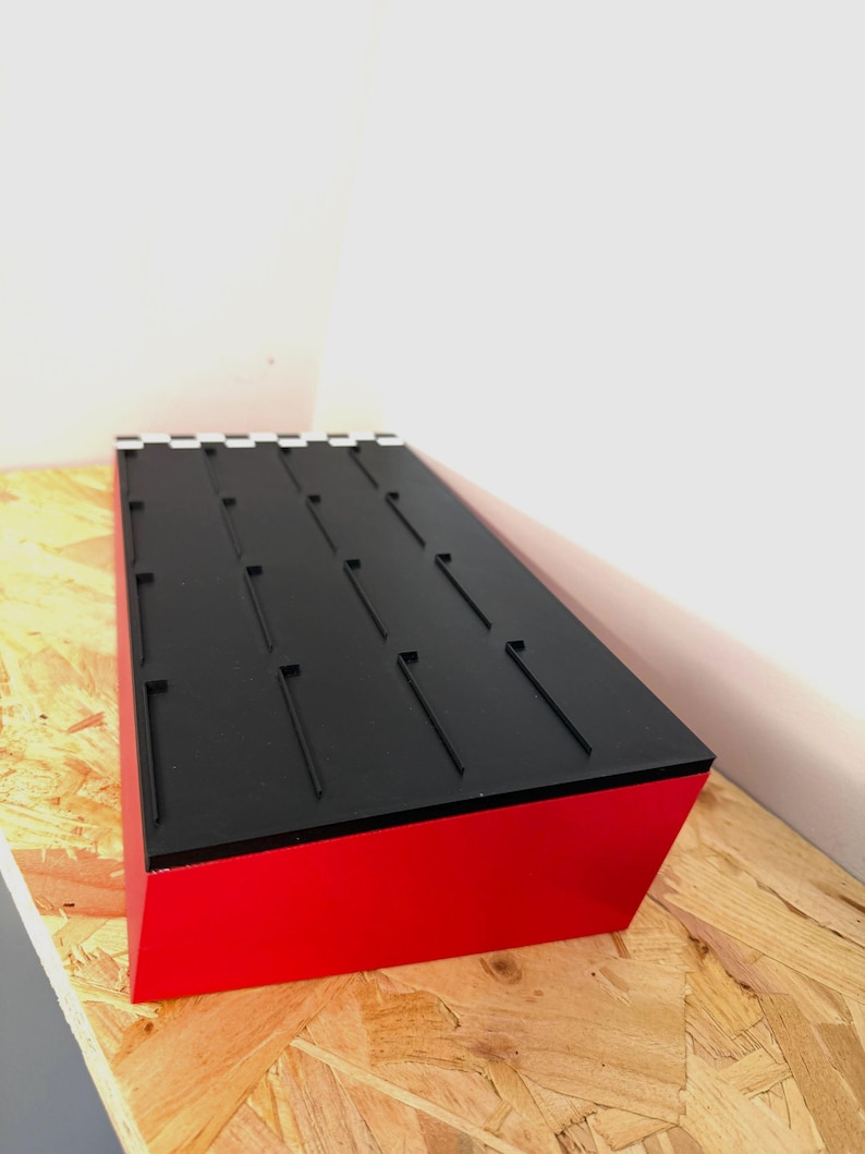 May include: A rectangular display stand with a red base and a black top. The top features several evenly spaced, vertical slots. The stand is angled, showcasing its design and construction.