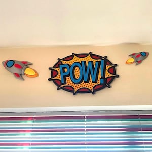 Puede incluir: Decoraci&oacute;n de pared con una burbuja de di&aacute;logo "POW!" estilo c&oacute;mic con texto azul y dos cohetes grises y rojos con llamas naranjas. Los art&iacute;culos est&aacute;n montados en una pared beige sobre una ventana con persianas coloridas.