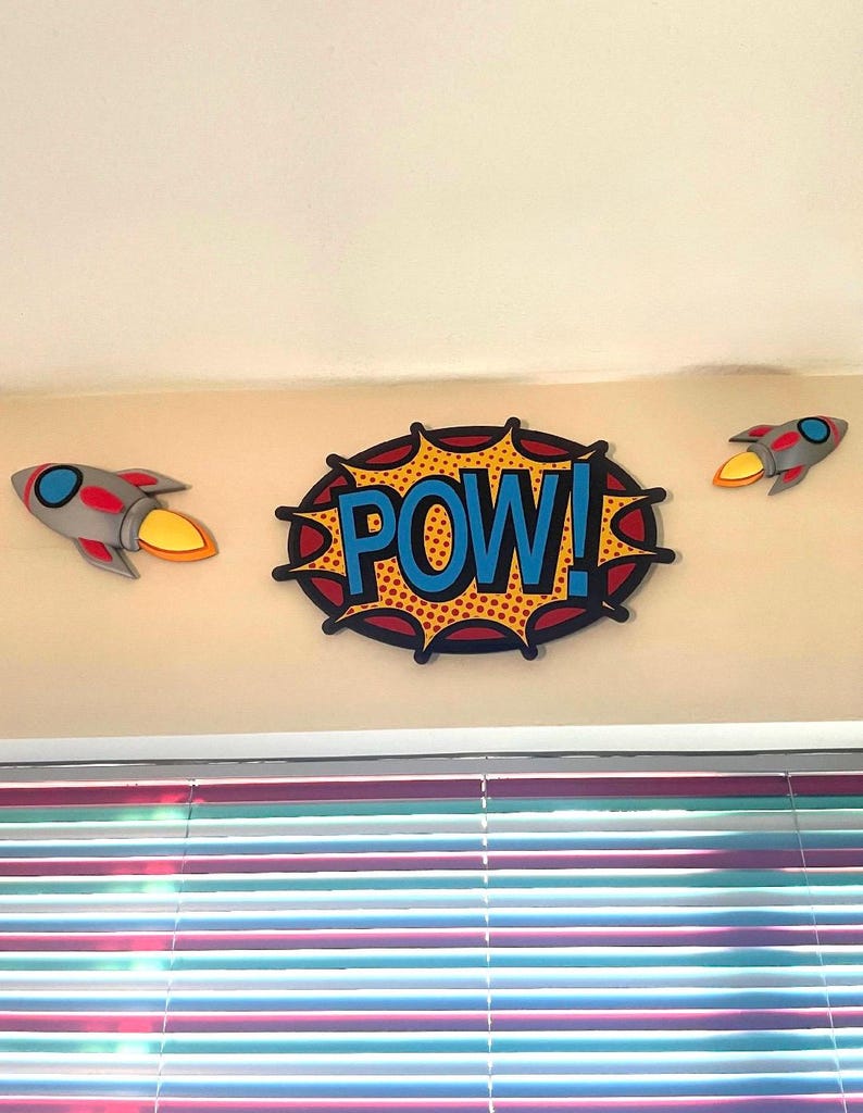 Puede incluir: Decoraci&oacute;n de pared con una burbuja de di&aacute;logo "POW!" estilo c&oacute;mic con texto azul y dos cohetes grises y rojos con llamas naranjas. Los art&iacute;culos est&aacute;n montados en una pared beige sobre una ventana con persianas coloridas.