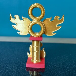 May include: A gold-coloured trophy with a flame and wheel design. The trophy has a red base and a cylindrical centre. The top features two circular elements and flame-shaped wings. The background is a solid blue.