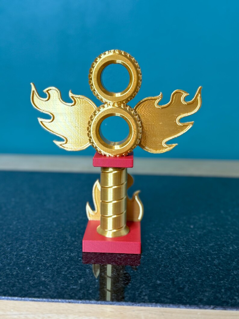 May include: A gold-coloured trophy with a flame and wheel design. The trophy has a red base and a cylindrical centre. The top features two circular elements and flame-shaped wings. The background is a solid blue.