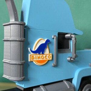 May include: A light blue toy truck with a silver exhaust pipe and fuel tank. The truck features a Dinoco logo with a blue dinosaur on a white and orange background. The truck is a toy.