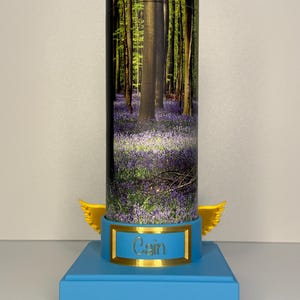 May include: A cylindrical container featuring a forest scene with purple flowers, mounted on a light blue square base. The base includes a gold-framed plaque with the name Cain and two small yellow wings extending from the sides of the cylinder.
