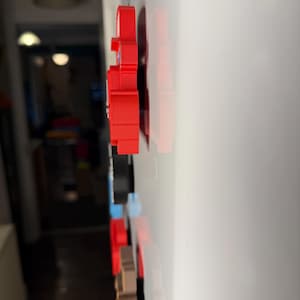 May include: A collection of colorful, 3D-printed magnets in various shapes, including a red bear, are attached to a white surface. The magnets are arranged in a vertical line, showcasing a range of colors and designs.