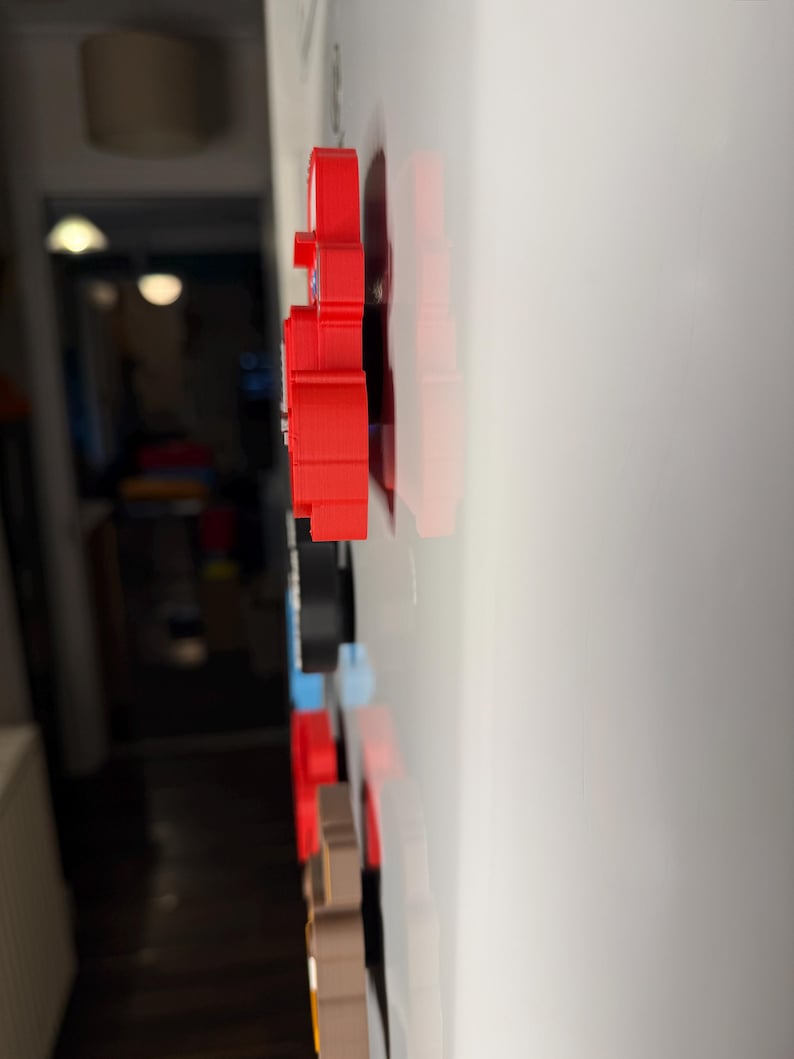 May include: A collection of colorful, 3D-printed magnets in various shapes, including a red bear, are attached to a white surface. The magnets are arranged in a vertical line, showcasing a range of colors and designs.