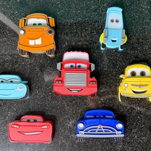 May include: A collection of colorful car-shaped magnets featuring characters from the animated movie "Cars." The magnets include a variety of car designs in shades of orange, blue, red, yellow, and blue, each with unique facial expressions.
