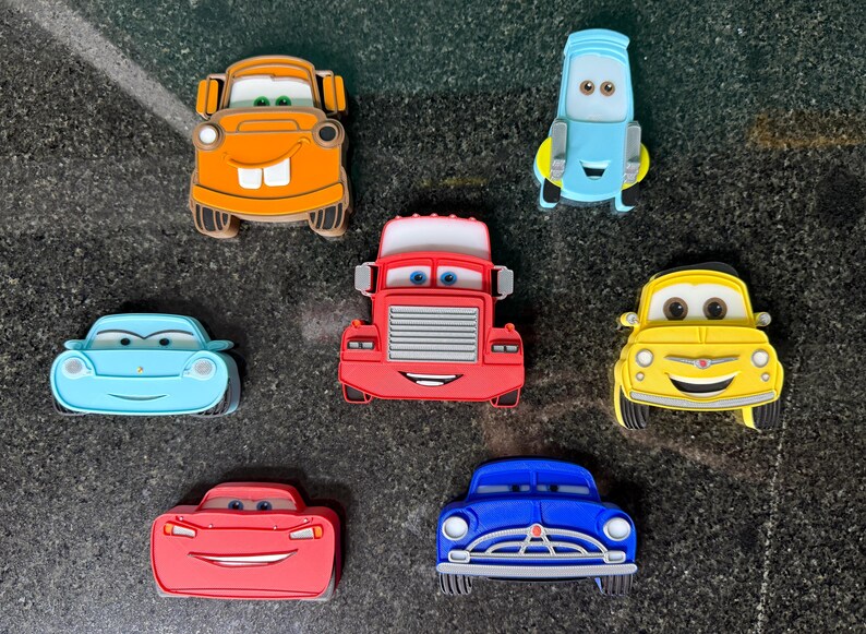 May include: A collection of colorful car-shaped magnets featuring characters from the animated movie "Cars." The magnets include a variety of car designs in shades of orange, blue, red, yellow, and blue, each with unique facial expressions.