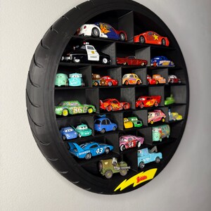 May include: A black, circular display shelf shaped like a tire, filled with various toy cars. The cars are of different colors and designs, arranged in individual compartments. A yellow lightning bolt graphic is on the bottom right.