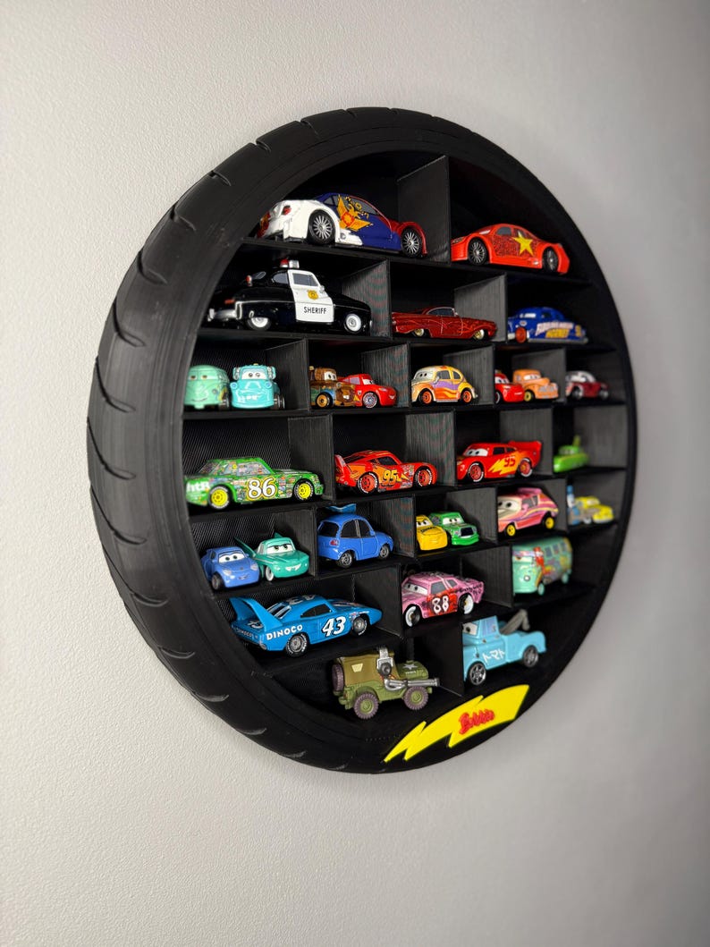 May include: A black, circular display shelf shaped like a tire, filled with various toy cars. The cars are of different colors and designs, arranged in individual compartments. A yellow lightning bolt graphic is on the bottom right.
