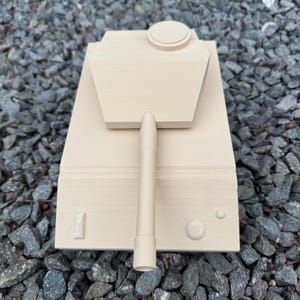 May include: A beige, monochromatic tank model featuring a central turret with a long, cylindrical barrel extending forward. The tank body includes a flat top surface with a circular hatch, a small rectangular detail, and two circular buttons.