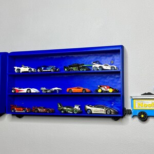 Two blue wall-mounted toy car display shelves connected to a customisable 3D-printed train engine labelled “Noah.” The shelves are filled with die-cast vehicles and designed to look like train carriages.