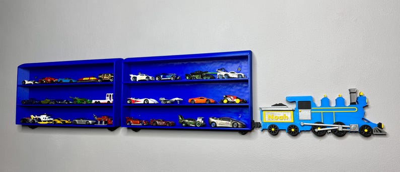 Two blue wall-mounted toy car display shelves connected to a customisable 3D-printed train engine labelled “Noah.” The shelves are filled with die-cast vehicles and designed to look like train carriages.