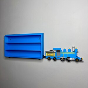 3D-printed toy train engine labelled “Noah” placed next to a matching blue toy car shelf, giving the illusion the train is pulling it. The shelf is empty and mounted on a grey wall.