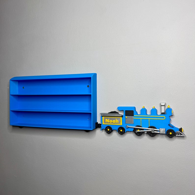 3D-printed toy train engine labelled “Noah” placed next to a matching blue toy car shelf, giving the illusion the train is pulling it. The shelf is empty and mounted on a grey wall.
