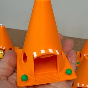 May include: Bright orange 3D-printed toy traffic cone with a white top and a small white door frame. The cone has a square base with a green ball on each side. The toy is a miniature replica of a traffic cone.