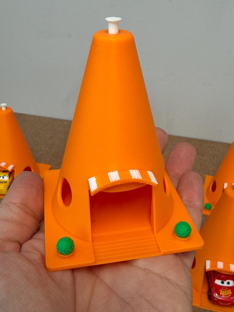 May include: Bright orange 3D-printed toy traffic cone with a white top and a small white door frame. The cone has a square base with a green ball on each side. The toy is a miniature replica of a traffic cone.