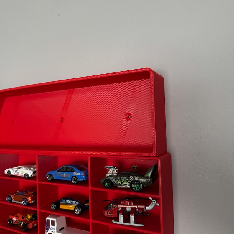 May include: Red plastic wall-mounted storage rack with multiple compartments for displaying toy cars. The rack is filled with various toy cars, including a blue car, a red car, a green car, and a helicopter.