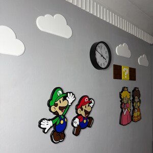 Op de afbeelding: Muurdecoratie met personages uit het Super Mario Bros. videospel. Inclusief Mario, Luigi, Princess Peach en Daisy, samen met witte wolkuitsparingen, een vraagtekenblok en een zwarte analoge klok.