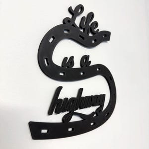 May include: Black wall art in the shape of a horseshoe with the words "Life is a highway" in a cursive font. The horseshoe has rectangular cutouts. The art is mounted on a white wall.