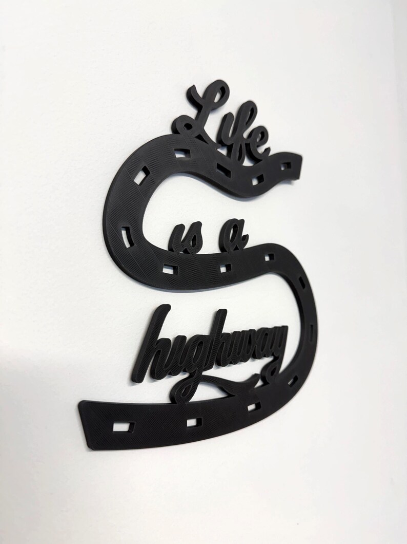 May include: Black wall art in the shape of a horseshoe with the words "Life is a highway" in a cursive font. The horseshoe has rectangular cutouts. The art is mounted on a white wall.