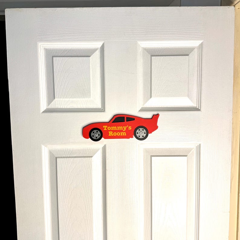 May include: A red car-shaped sign with the text "Tommy's Room" in yellow lettering. The sign is affixed to a white door with rectangular paneling. The car has black wheels and a spoiler.