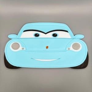 May include: A light blue car-shaped placemat featuring a cartoon-style front view with large white eyes, black pupils, and a small colorful emblem on the hood. The design includes two circular headlights with silver mesh details and black tires.