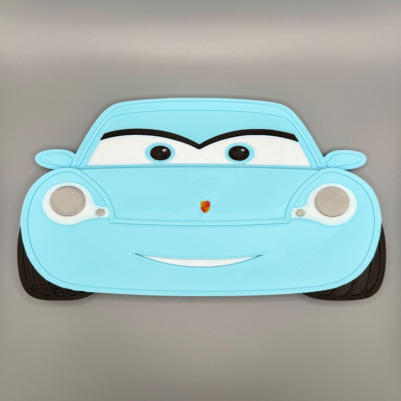 May include: A light blue car-shaped placemat featuring a cartoon-style front view with large white eyes, black pupils, and a small colorful emblem on the hood. The design includes two circular headlights with silver mesh details and black tires.