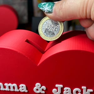 Hand placing a coin in a Love heart-shaped piggy bank, ideal for saving coins, Valentine’s Day gifts, or romantic decor. A unique 3D-printed coin bank for kids or couples, perfect as a gift, decorative piece, or practical savings tool.