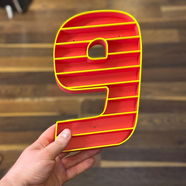 May include: A red number nine-shaped shelf with yellow trim. The shelf has horizontal dividers, creating multiple storage spaces. The number is held in a hand against a wooden background.