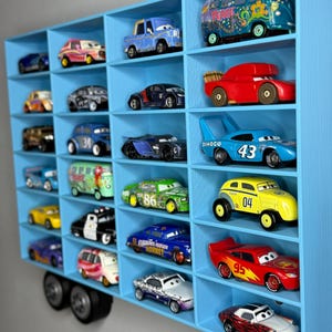 May include: A blue, wall-mounted display shelf filled with various toy cars. The shelf has multiple compartments, each holding a different car model. The cars are in various colors, including red, blue, yellow, and green. The shelf is designed to resemble a truck.