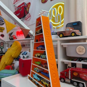 May include: An orange and white shelf unit shaped like a triangle, with the text "COZY CONE MOTEL" on the side. The shelves display a collection of toy cars. The background includes a red Lightning McQueen hat and a "Lightning" banner.