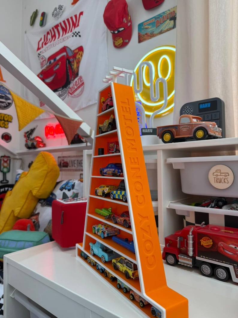 May include: An orange and white shelf unit shaped like a triangle, with the text "COZY CONE MOTEL" on the side. The shelves display a collection of toy cars. The background includes a red Lightning McQueen hat and a "Lightning" banner.